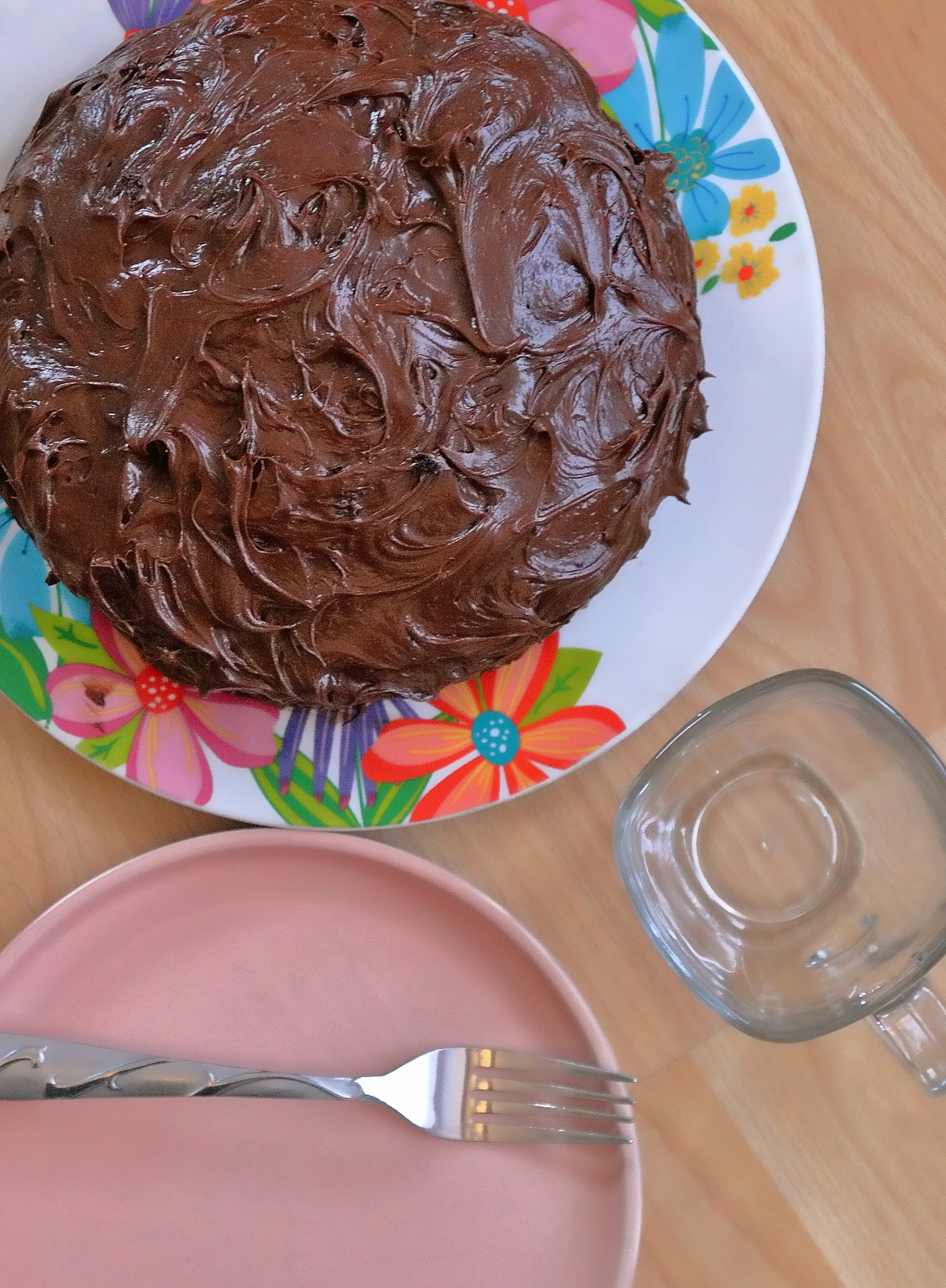 Sjokoladekake med strukturert frosting på en blomsterplate, ved siden av et tomt glasskrus og en rosa tallerken med en gaffel