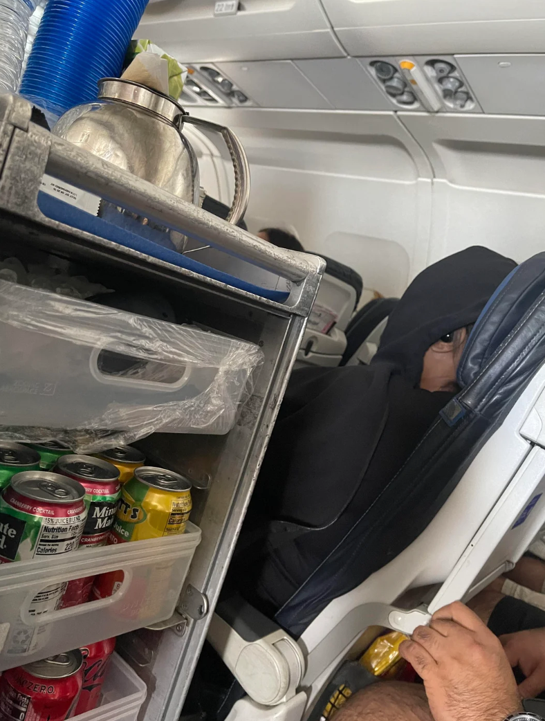 A idiosyncratic   successful  a achromatic  hoodie peers retired  portion    seated connected  a plane, adjacent  to a stocked beverage cart