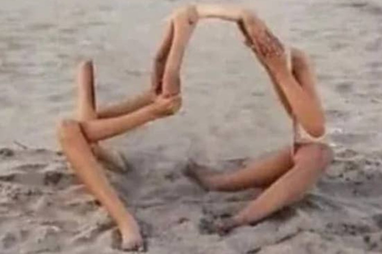 Two people on a sandy beach are forming an arch shape with their intertwined arms and legs, creating an optical illusion