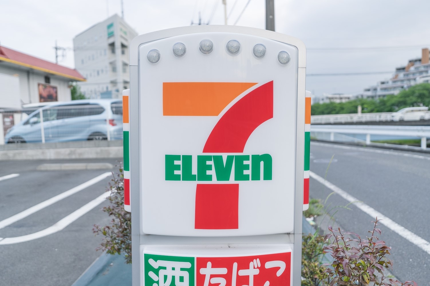 セブンイレブンの看板が駐車場に立っている。背景には道路と建物が見える。