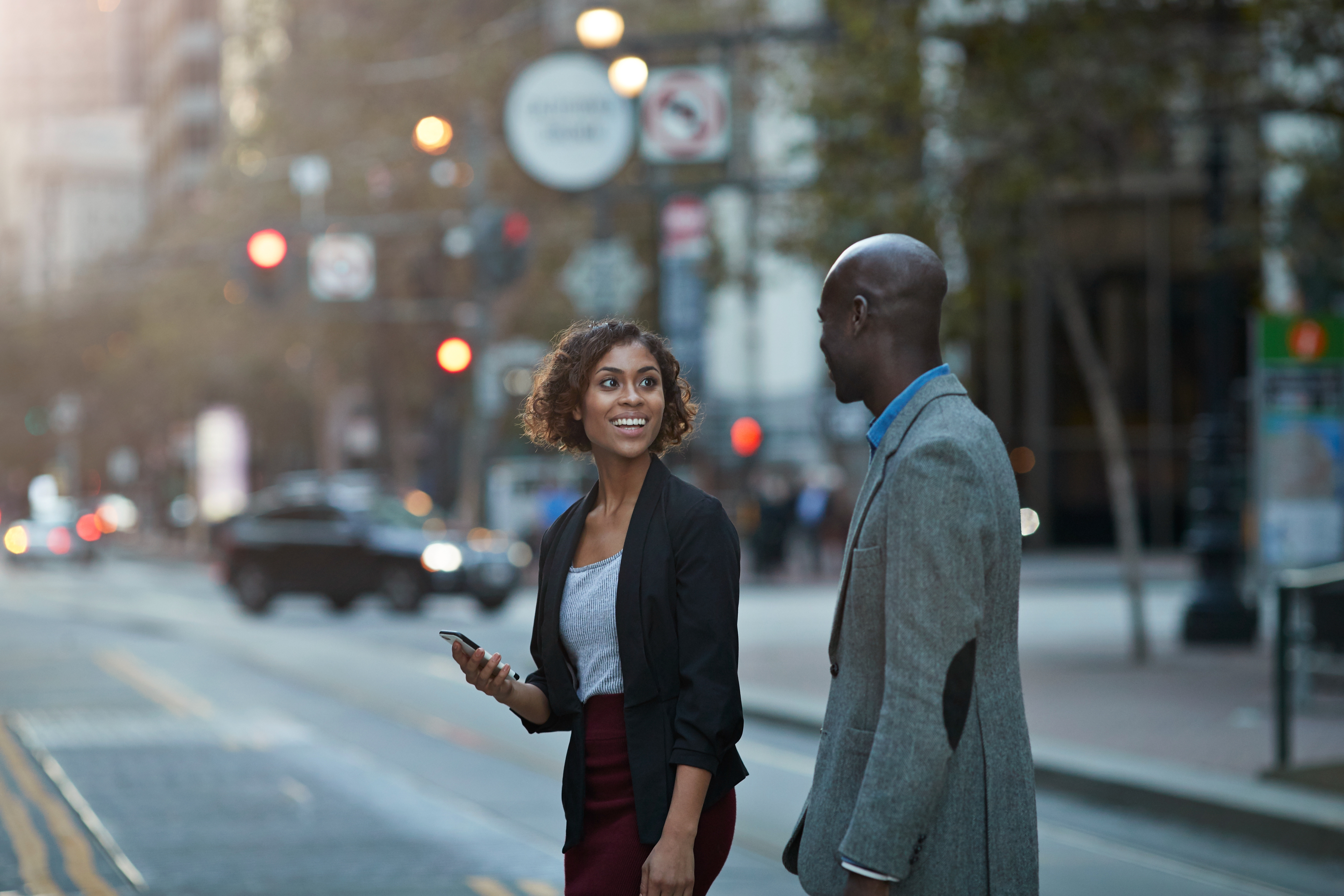 Two people in business attire are talking and walking across a city street. One is holding a smartphone and smiling