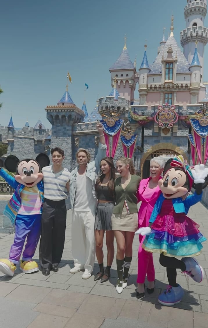 Five people pose with Mickey and Minnie Mouse in front of a castle. Two wear casual outfits, while others are in trendy and brightly colored suits