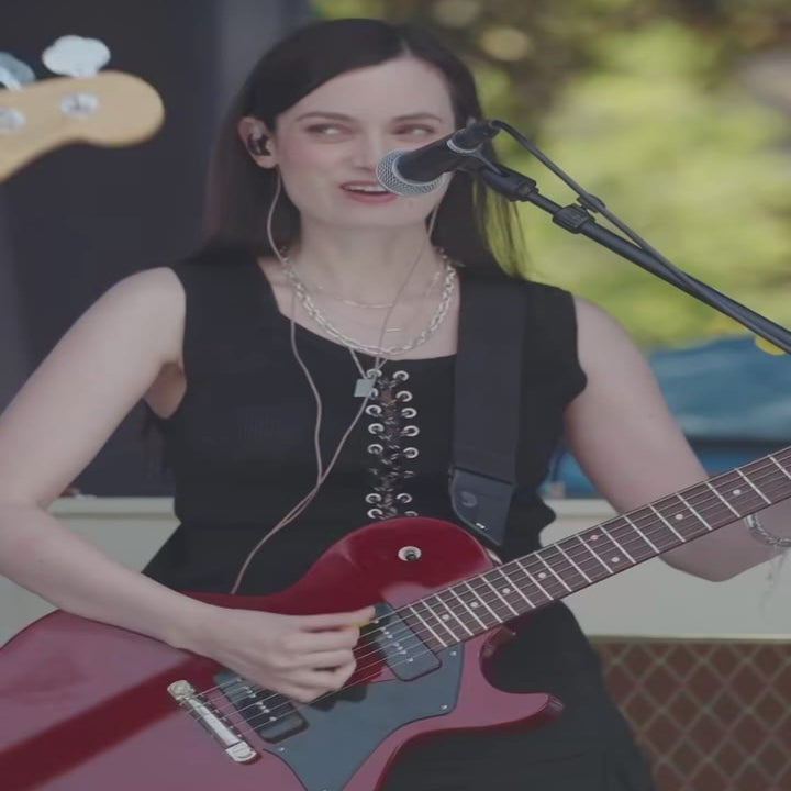 Musician performs on stage with a guitar, wearing a sleeveless dress and headphones