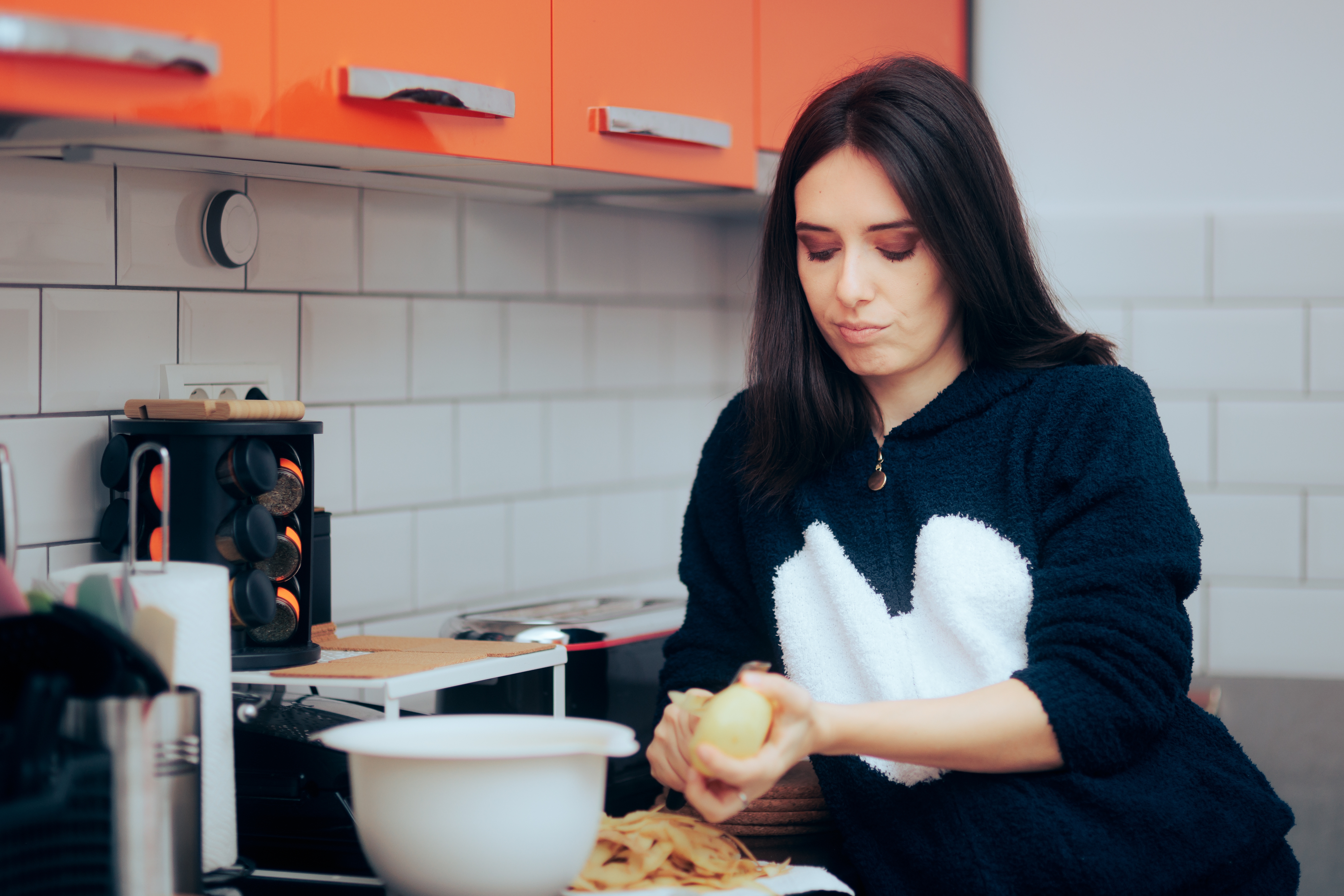 Kvinne som skreller poteter på et kjøkken med moderne skap, iført en genser med et stort hjerteutforming. Hun virker fokusert