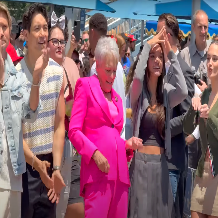A group of people celebrate outdoors. A person in a bright suit stands out, smiling and dancing among casually dressed others