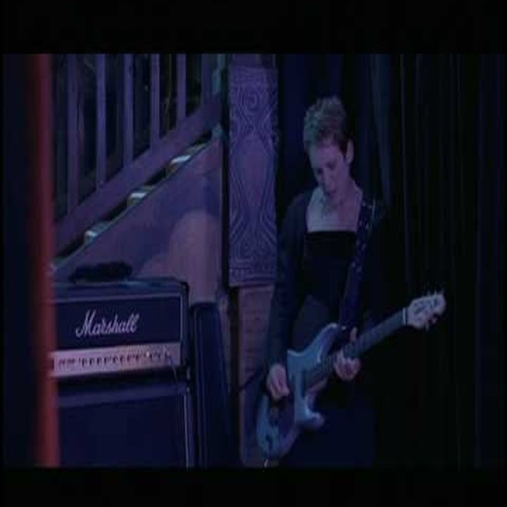 A musician plays electric guitar beside a large amplifier on a dimly lit stage