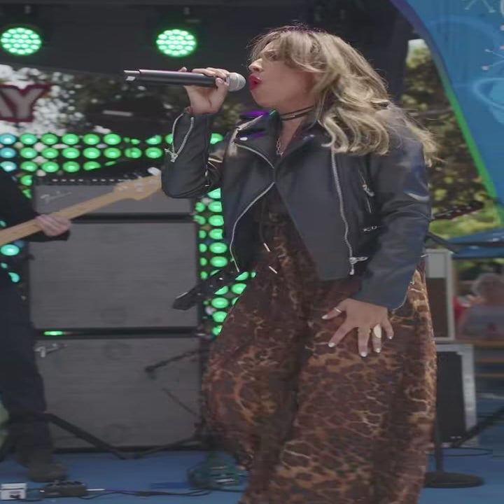 Person singing on stage in a flowy animal print dress and leather jacket, holding a microphone with a vibrant background