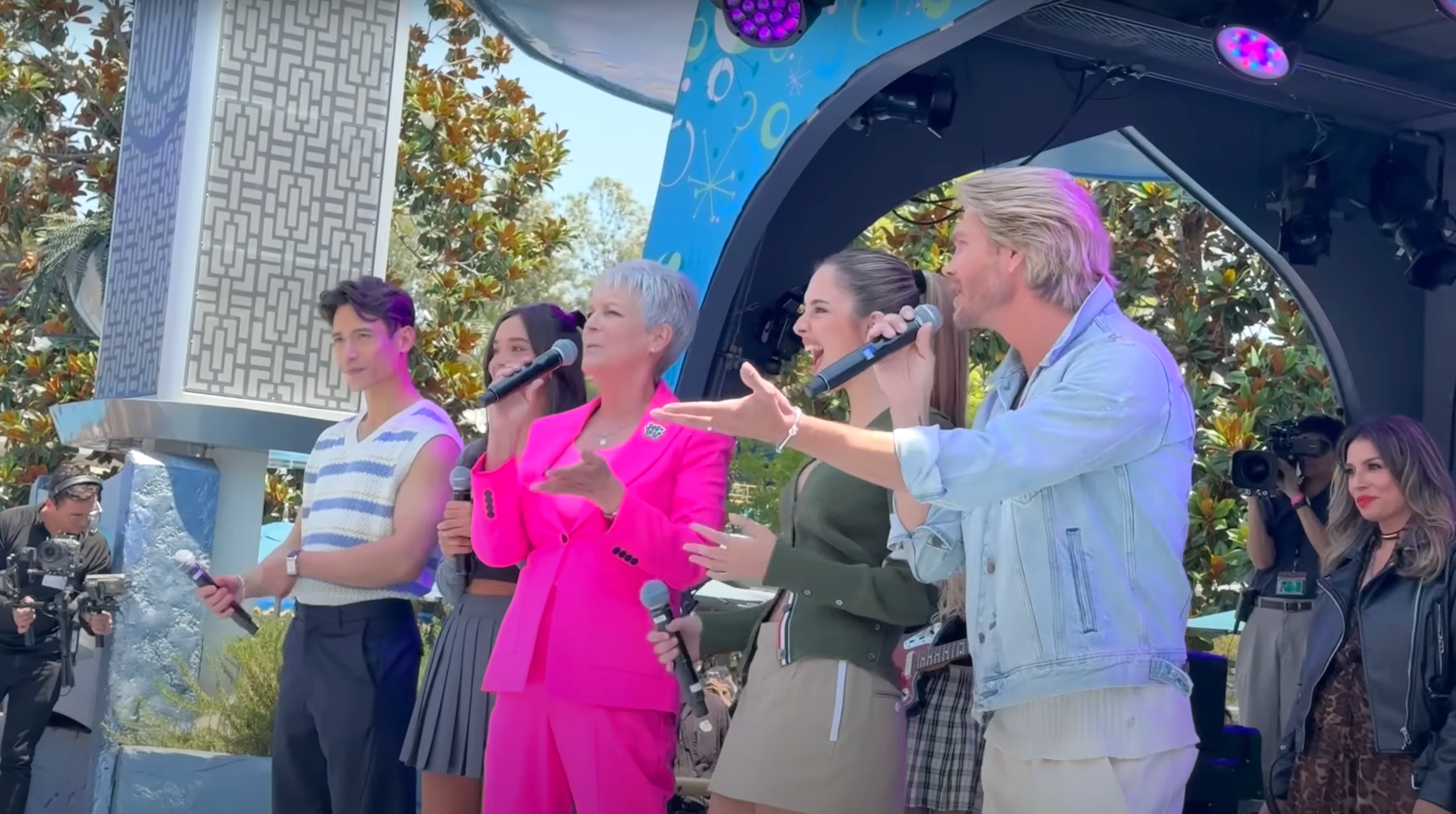 A group performs on stage. The person in the center is wearing a bright suit and holding a microphone. Others join in singing and engaging the audience