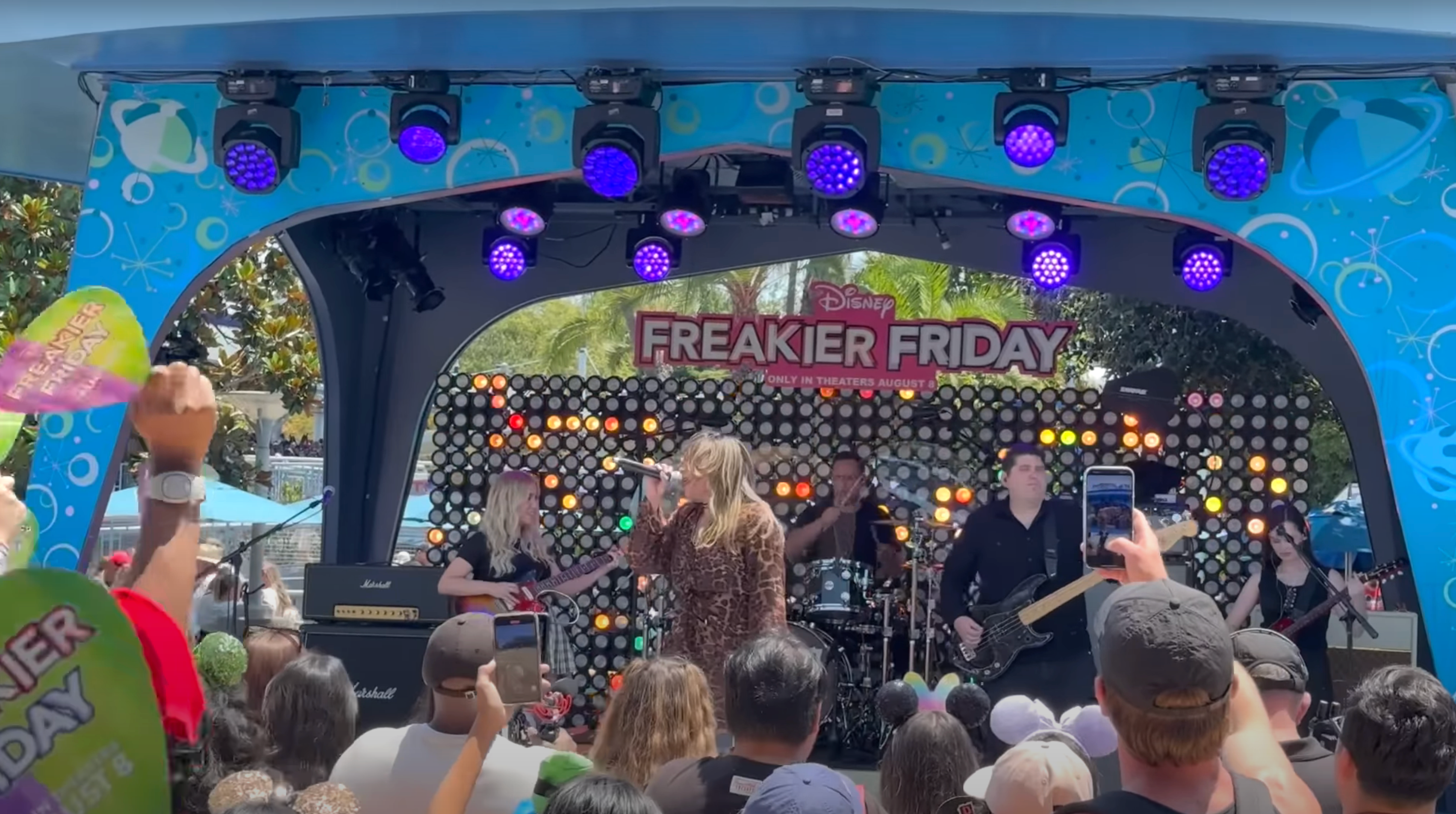 Band performing on stage at Disney's Freakier Friday event with a large crowd watching. A sign displaying the event name is visible in the background
