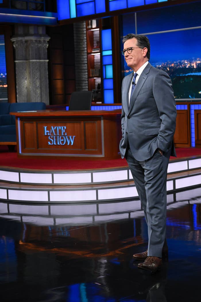 Stephen Colbert in a suit stands on a talk show set featuring a desk and large video screen backdrop