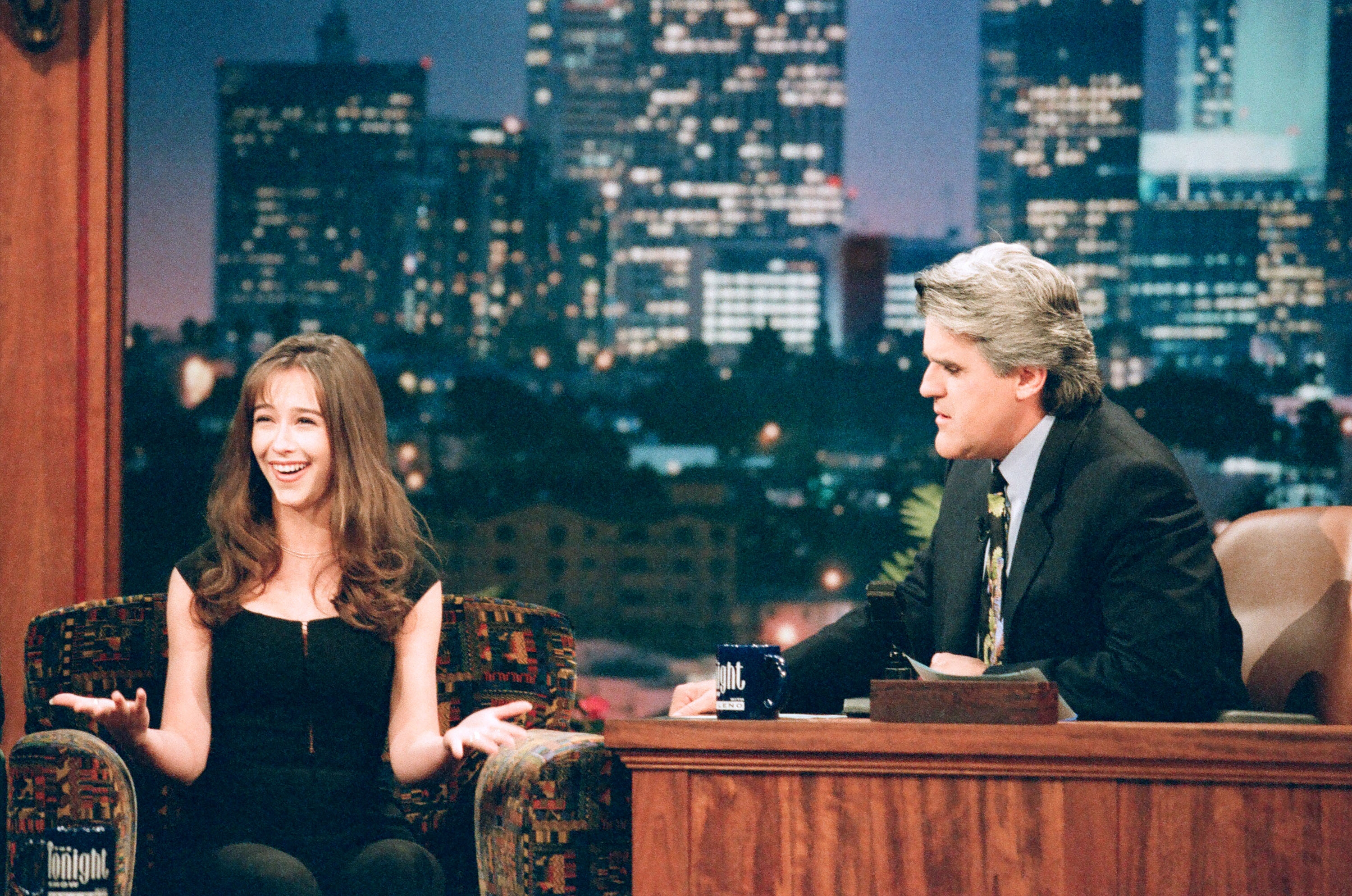 A guest in a black outfit chats animatedly with a talk show host, seated next to her, on a set with a cityscape backdrop