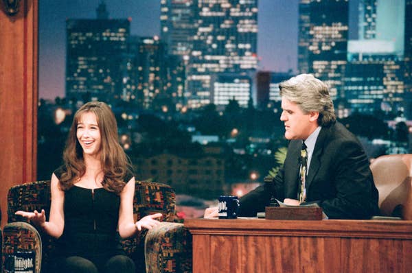 A guest in a black outfit chats animatedly with a talk show host, seated next to her, on a set with a cityscape backdrop