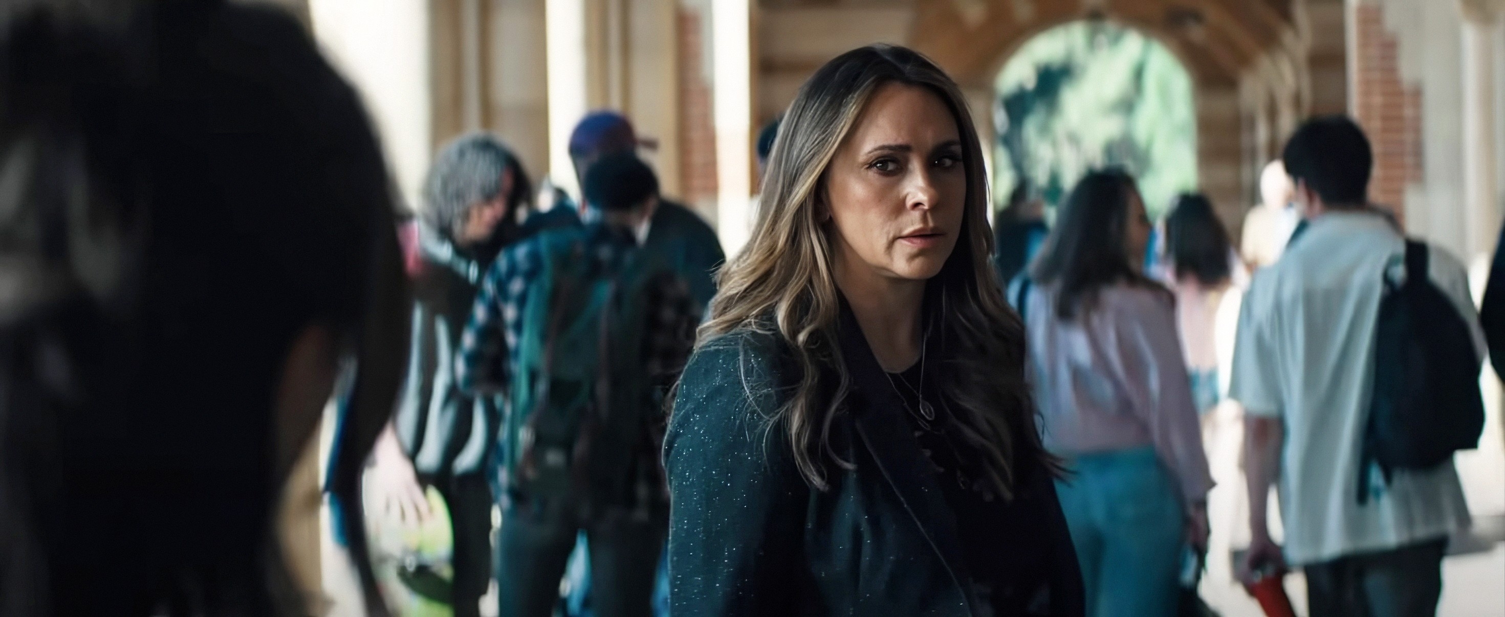 Woman in a crowd, looking concerned, with flowing hair and a stylish jacket, possibly a scene from a movie or TV show