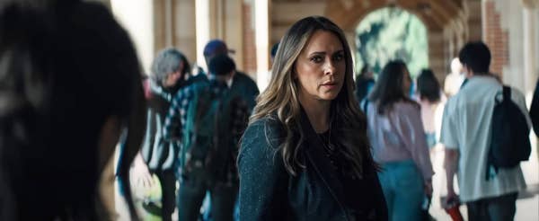 Woman in a crowd, looking concerned, with flowing hair and a stylish jacket, possibly a scene from a movie or TV show