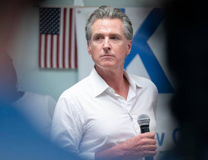 A person holding a microphone looks to the side at an event, with a U.S. flag in the background