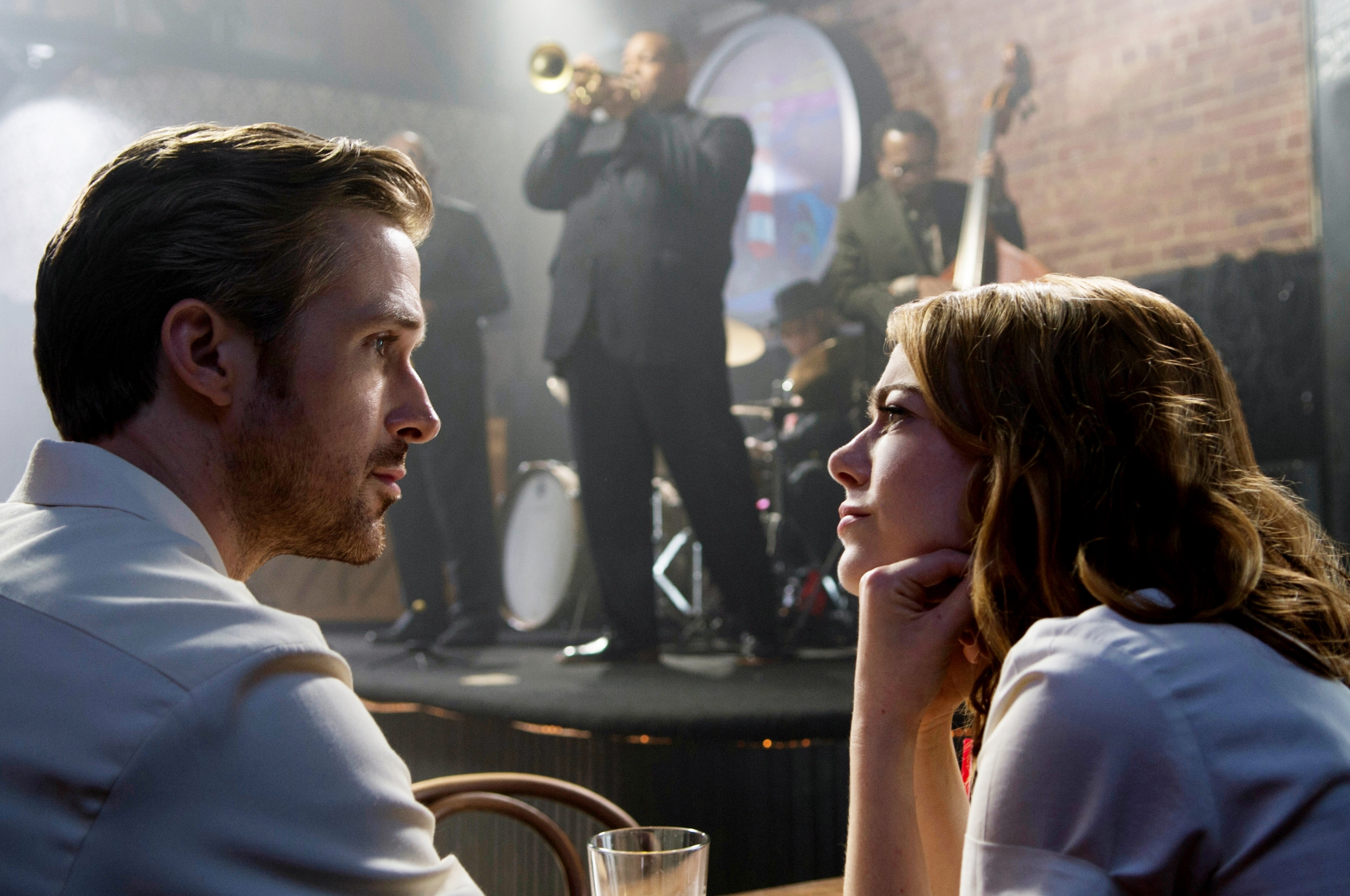 Ryan Gosling and Emma Stone gazing at each other in a dimly lit jazz club, with a band playing instruments in the background as Sebastian and Mia in La La Land