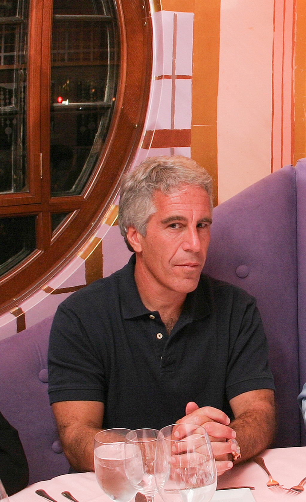 A man with gray hair, wearing a dark polo shirt, sits at a dining table with his hands clasped, surrounded by empty glasses