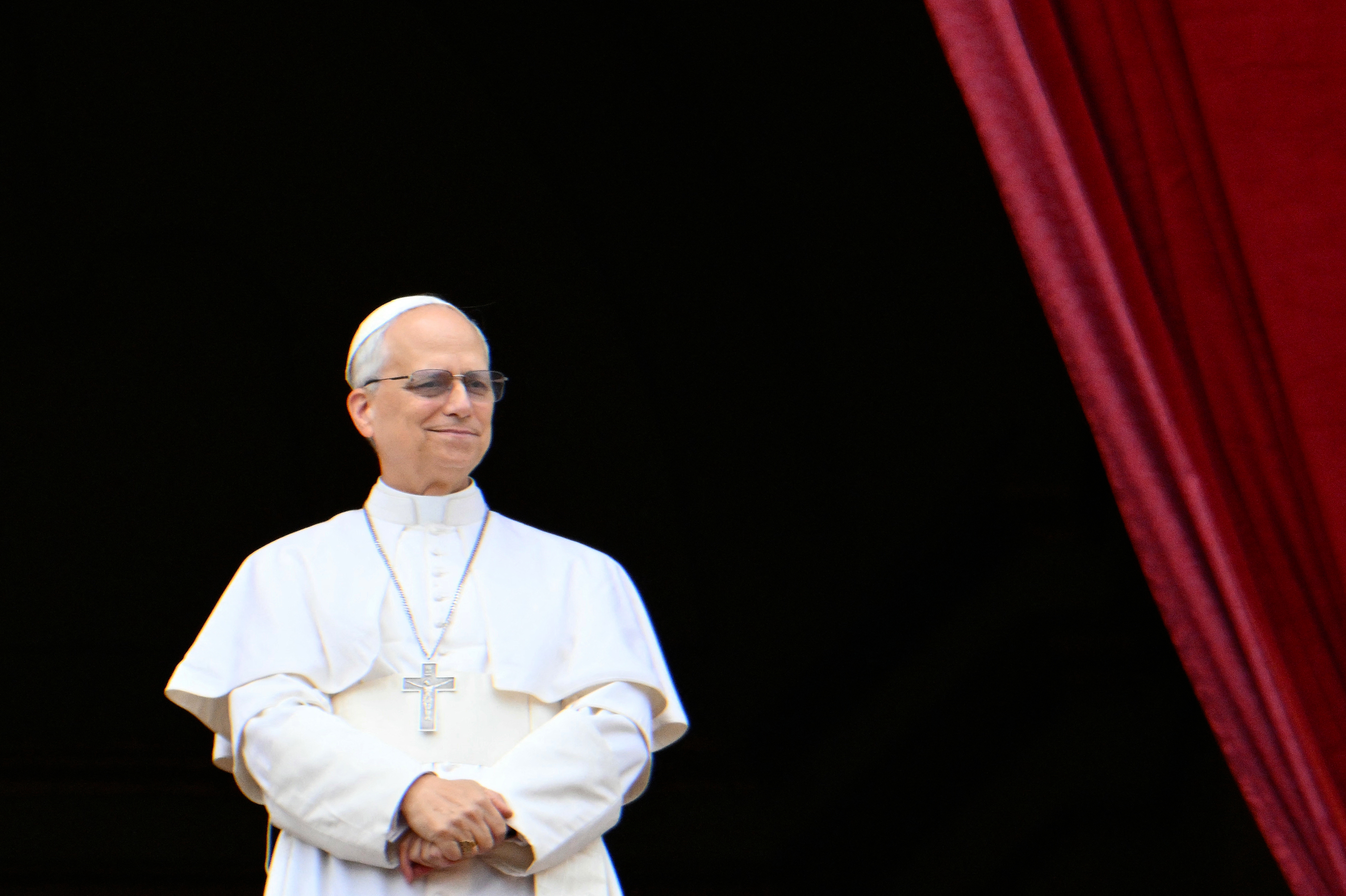 A man dressed as the Pope stands against a dark backdrop, wearing traditional papal attire, including a cross necklace