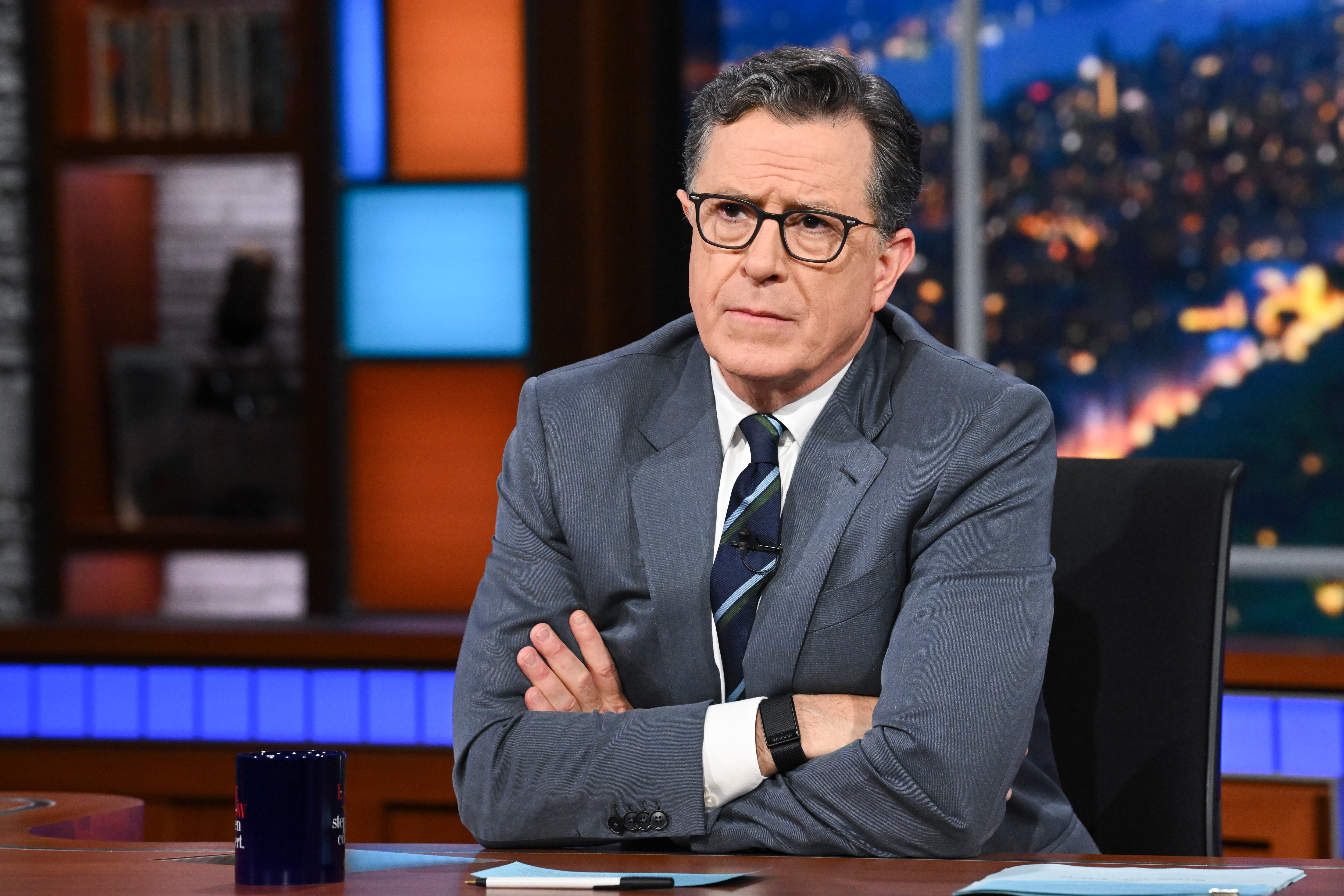 TV host sits at a desk, arms crossed, wearing a suit and tie, with a thoughtful expression