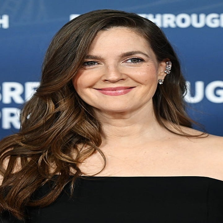 Actor with long, wavy hair smiles in an off-the-shoulder black dress at a "Breakthrough Prize" event backdrop