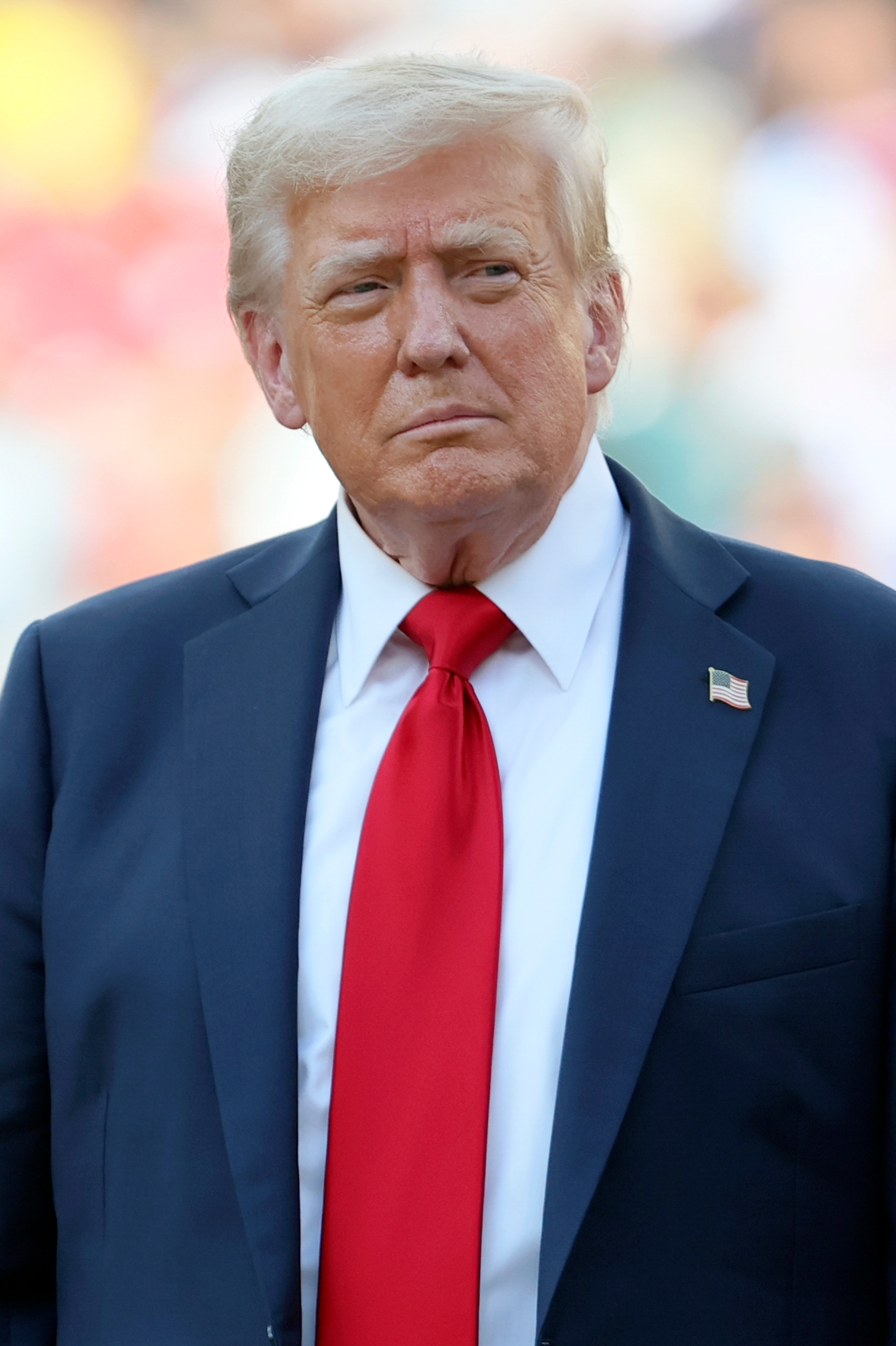 A man in a suit with a red tie stands, looking serious, against a blurred background