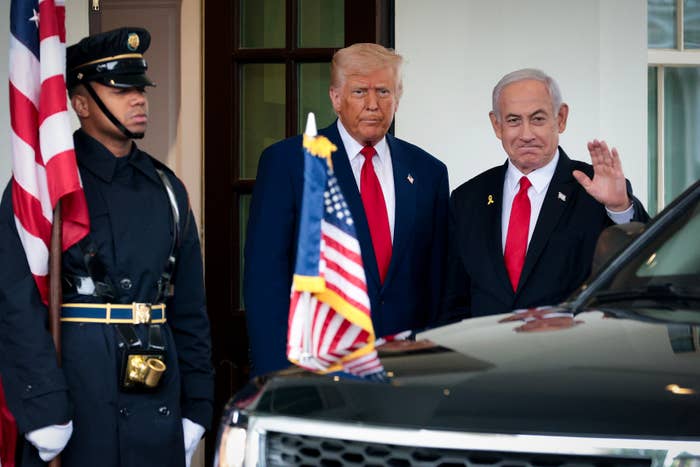 Two men in business suits stand near a car with American flags, one waving. A ceremonial guard is in uniform beside them