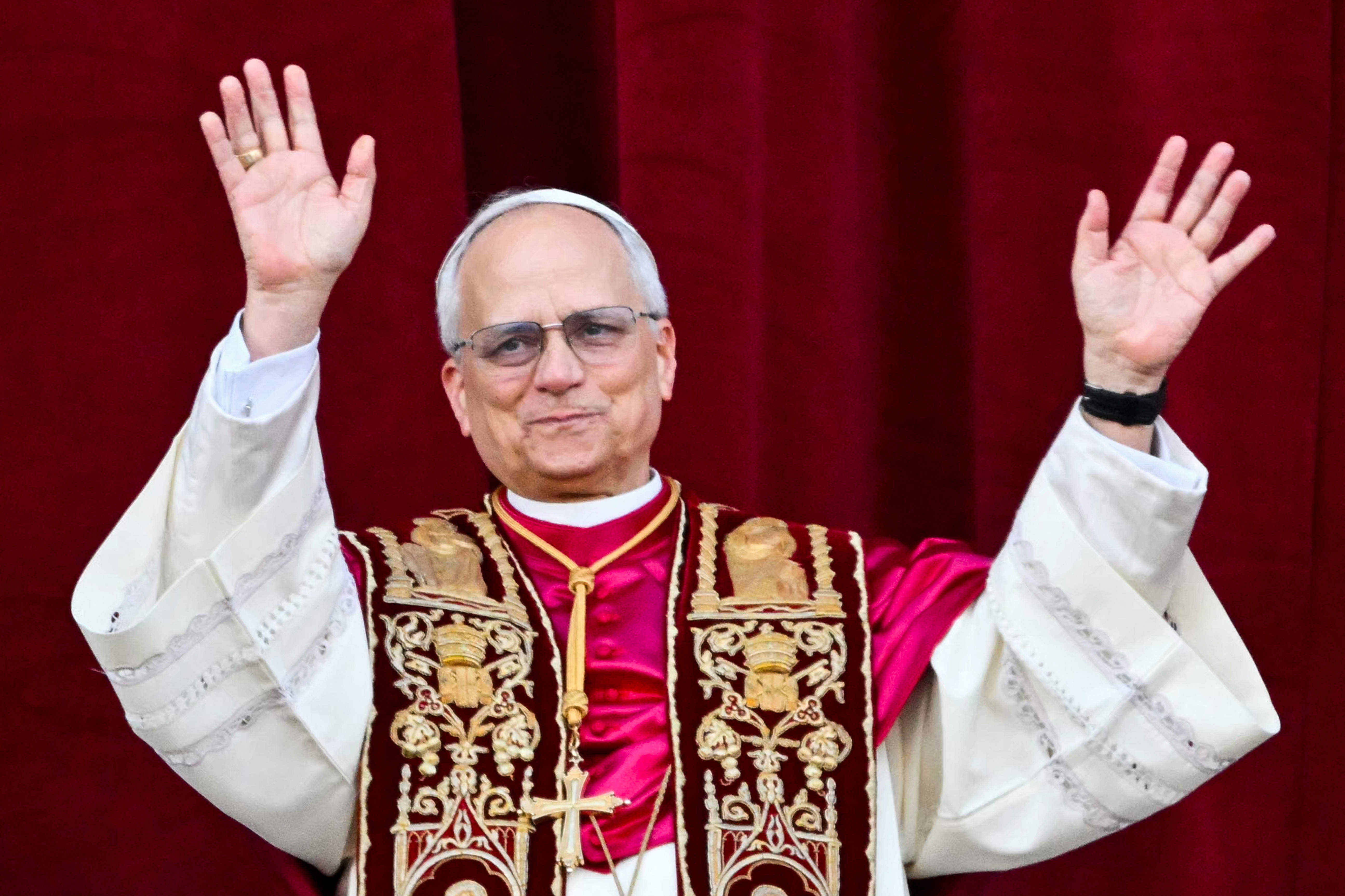 A man in religious attire gestures with both hands raised in front of a textured backdrop