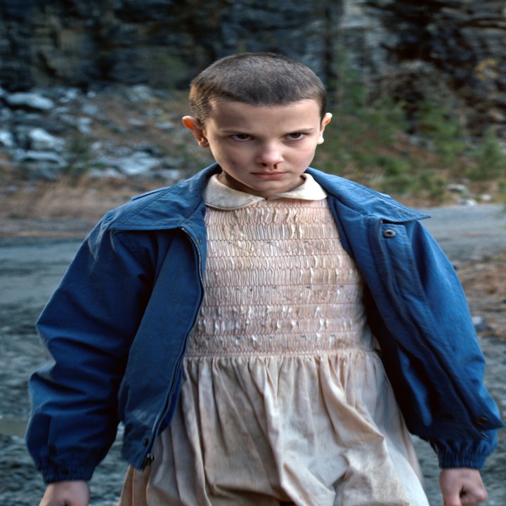 Person in blue jacket over a beige dress stands outdoors on a dirt path, looking focused