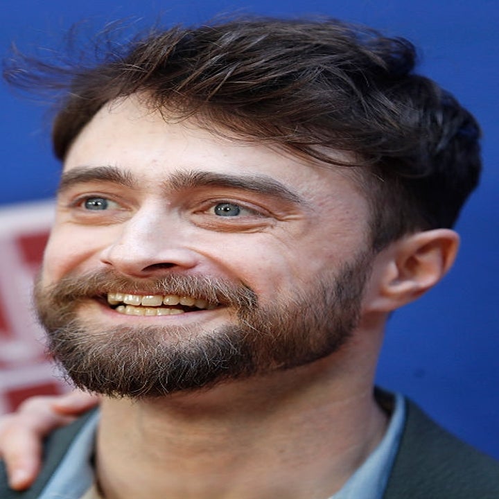 Person smiling at an event, wearing a suit with an open-collared shirt. Background features a blurred red and white pattern