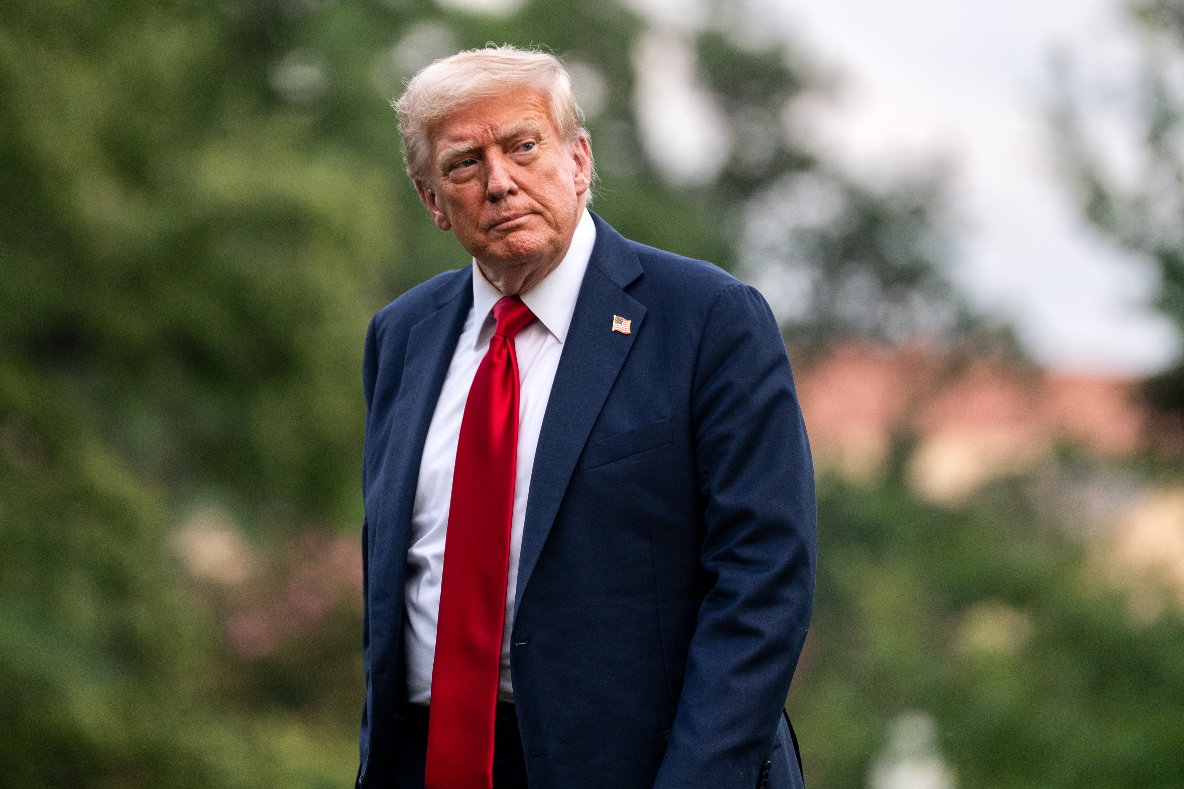 A person in a suit and red tie is outside, looking to the side, with a blurred backdrop of greenery