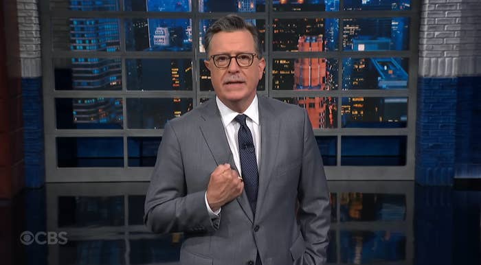 Stephen Colbert in a suit and tie is standing in a studio with a cityscape backdrop, gesturing with his hand while speaking
