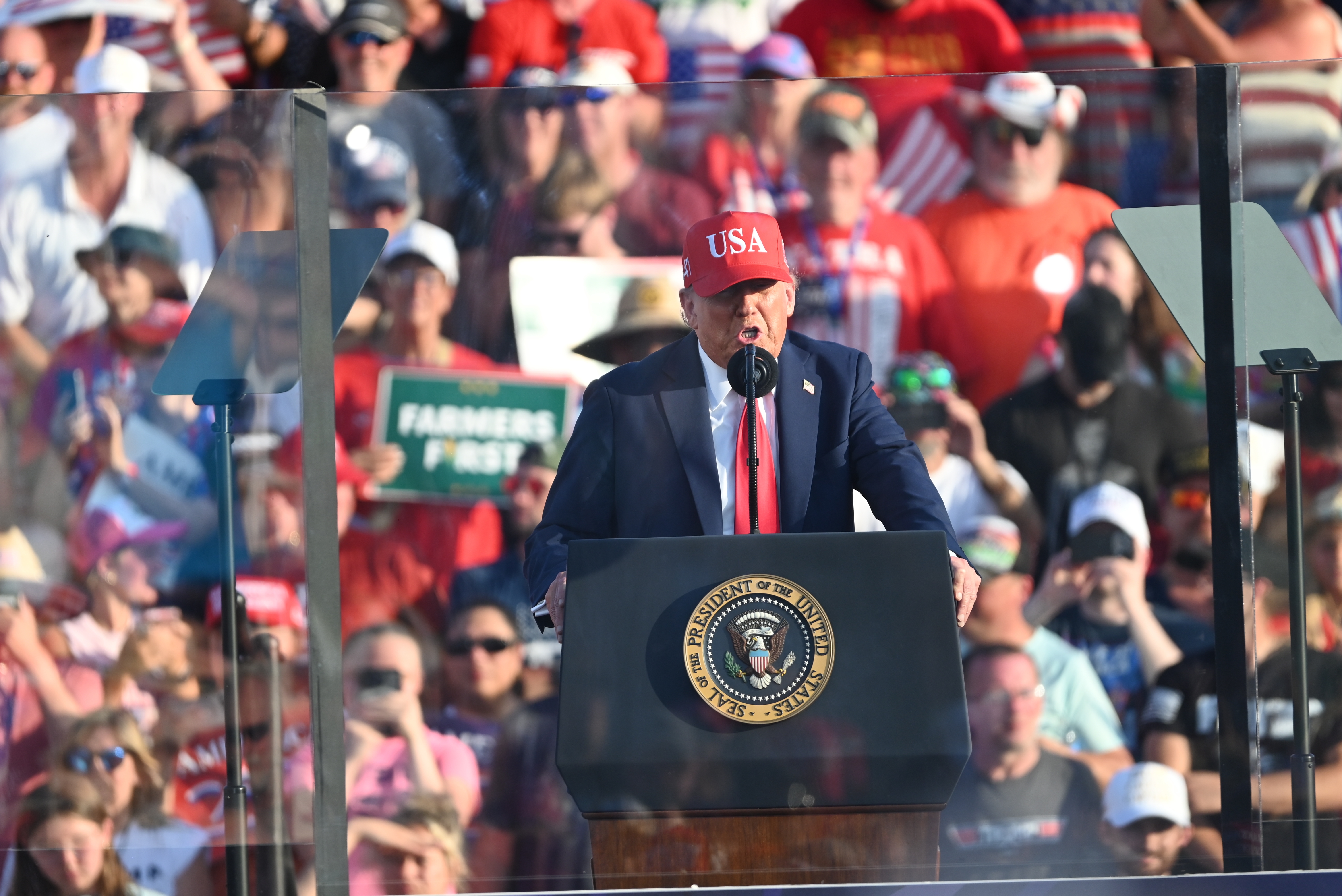 Donald Trump in a suit and a &quot;USA&quot; hat speaks at a podium surrounded by a crowd holding signs at a public event