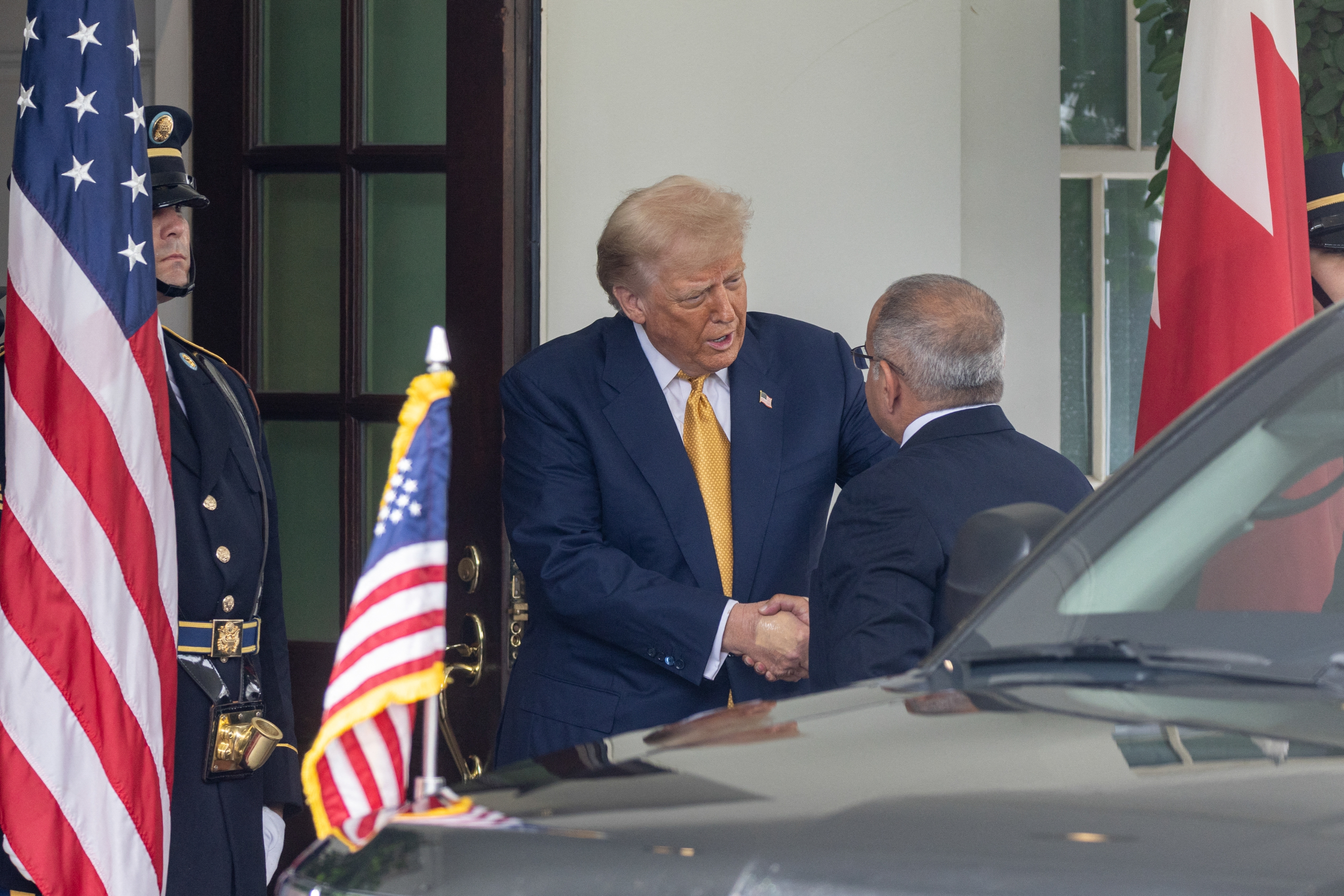 Two men shake hands outside a building with flags and guards. One wears a suit and tie, appearing formal and official