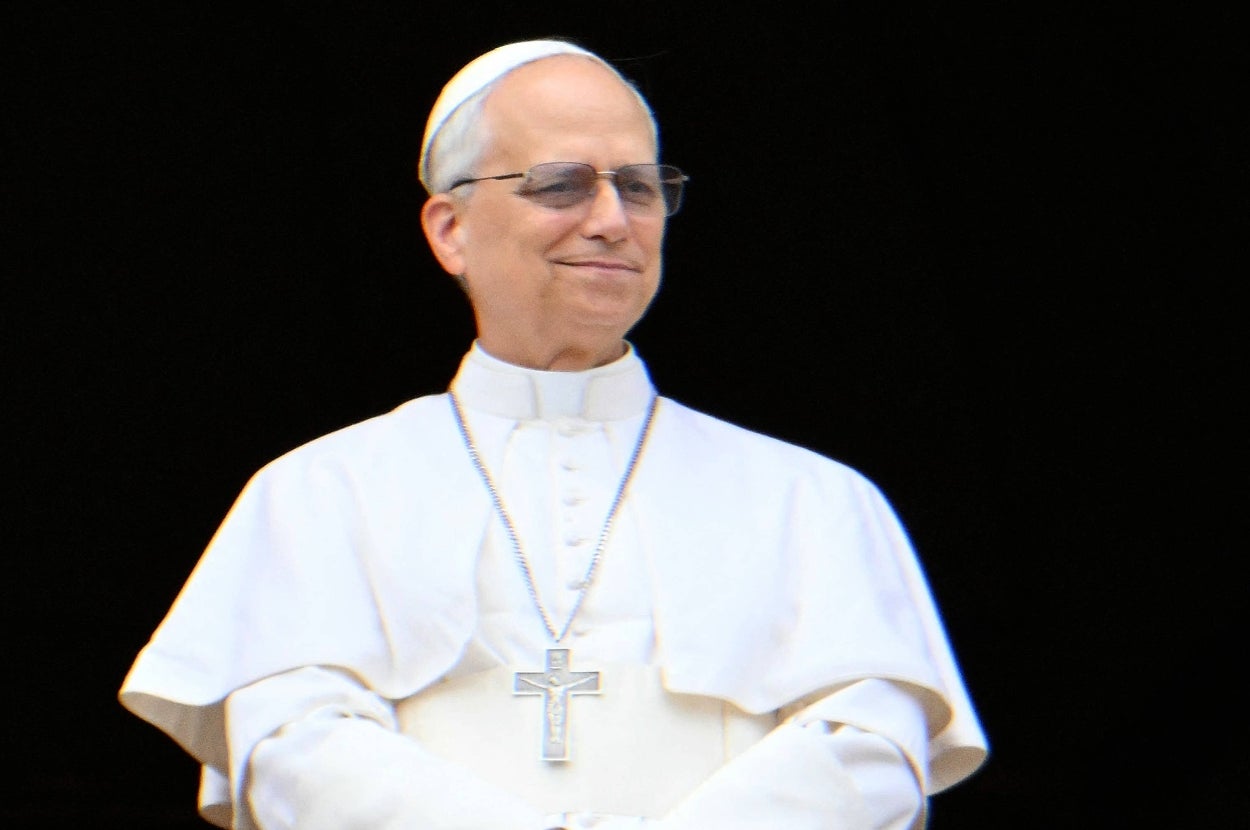A man dressed in traditional papal attire with glasses and a cross necklace stands confidently, smiling