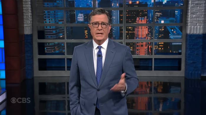 A man in a suit gestures while speaking on a talk show set with a cityscape backdrop