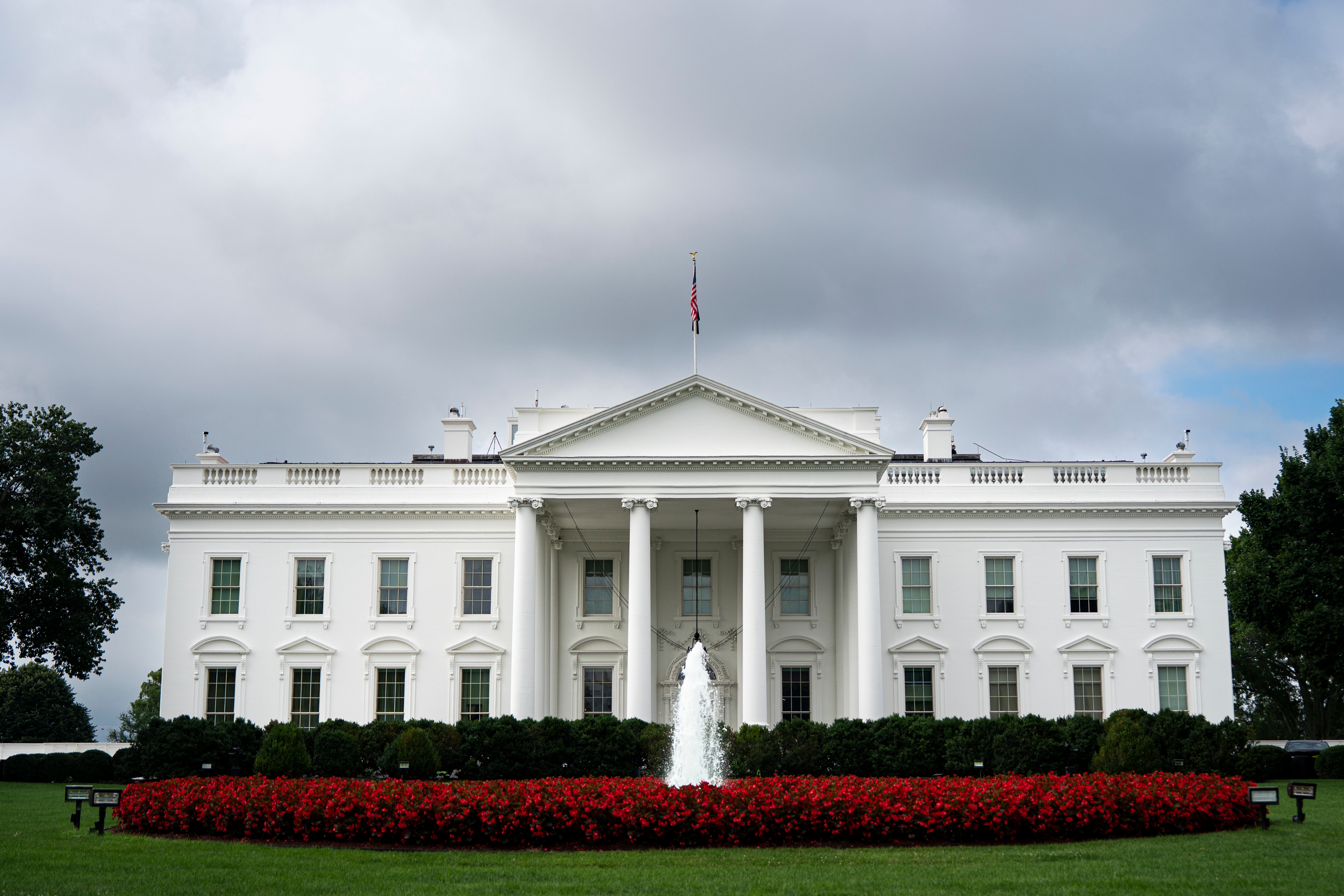 White House with fountain in front, surrounded by greenery and red flowers, under a cloudy sky