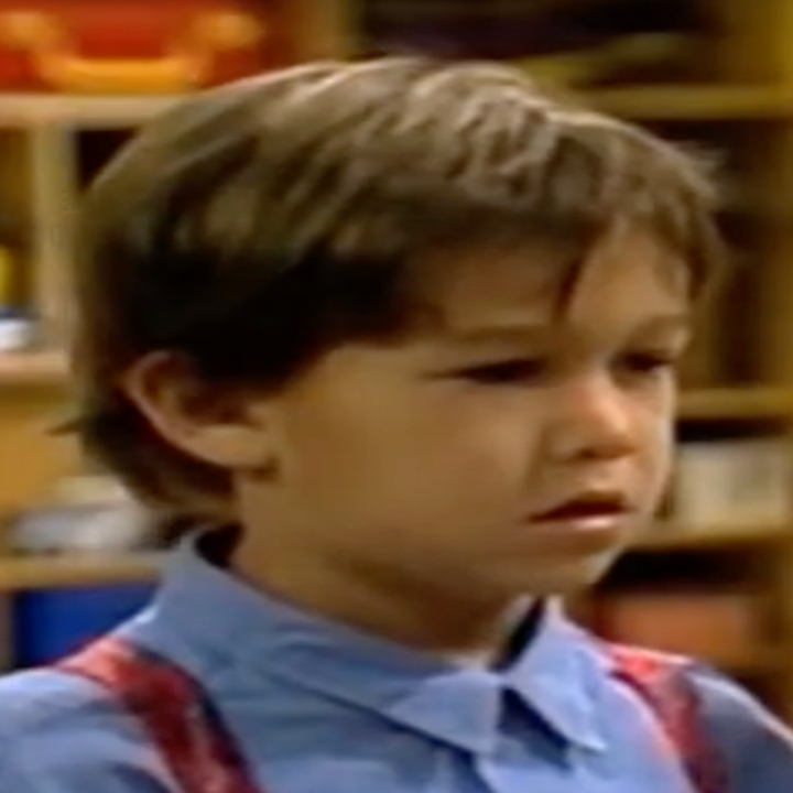 Young child in a blue shirt and red suspenders, standing indoors with a concerned expression
