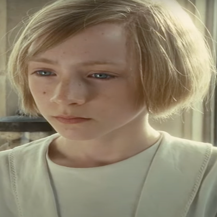 Young person with short, light hair stands in front of a window, wearing a simple light shirt, looking pensively into the distance