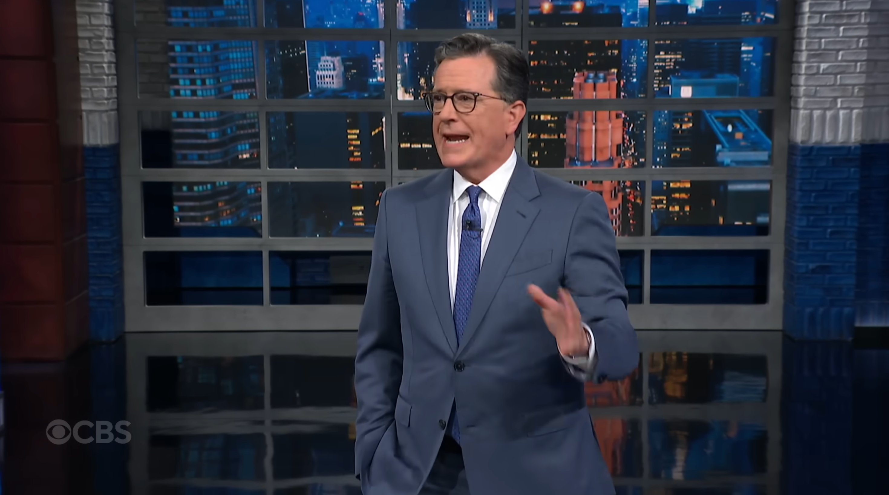 A person in a suit gestures while speaking on a talk show set with a cityscape backdrop