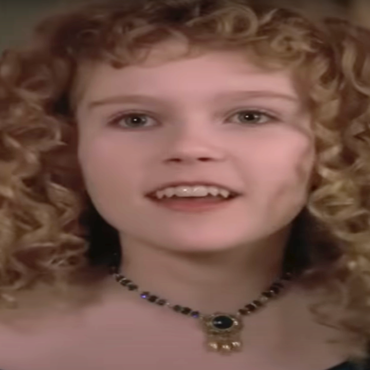 Person with curly hair wears a period-style dress and necklace, smiling and looking upward