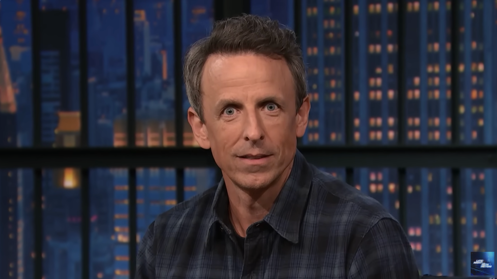 Seth Meyers on a late-night talk show set, wearing a plaid shirt, looking towards the camera with a city skyline backdrop