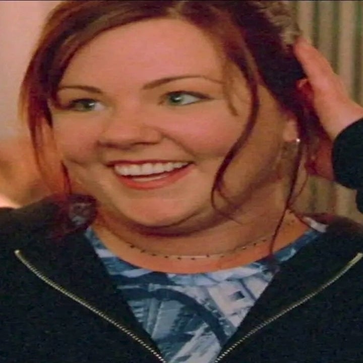 Person smiling warmly, wearing a casual black zip-up hoodie over a patterned top, in a cozy interior setting with soft lighting