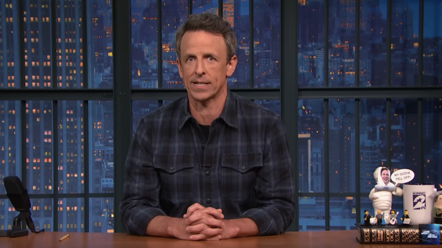 Seth Meyers sits at a desk in a TV studio, wearing a casual plaid shirt, with cityscape backdrop visible through large windows