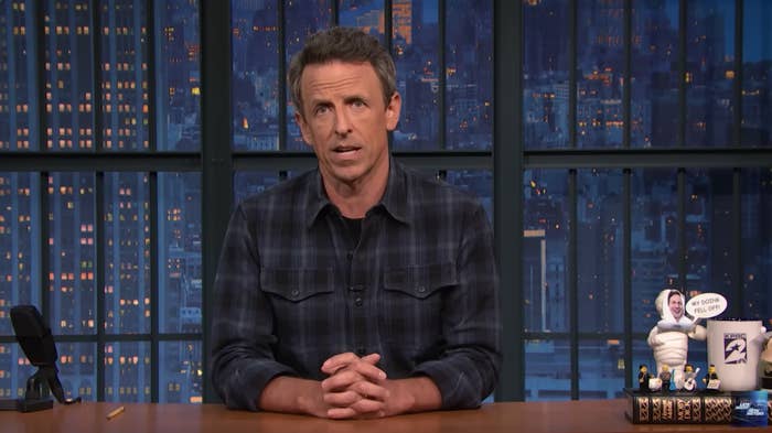 Seth Meyers sits at a desk with city lights visible through a window backdrop, speaking during a late-night show segment