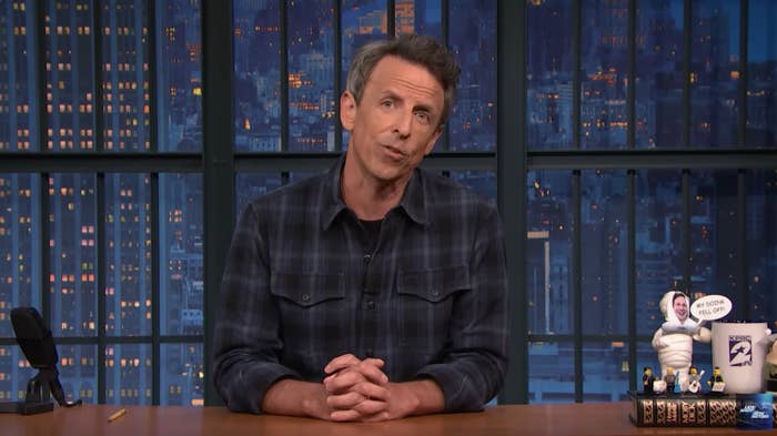 Seth Meyers in a plaid shirt speaks while seated at a desk on a city skyline set, with playful figurines beside him