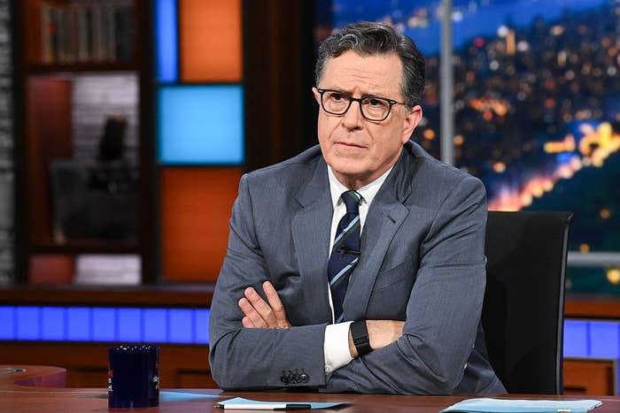 Man in a suit seated at a TV late-night show desk, arms crossed, with a serious expression, set against an urban nightscape backdrop
