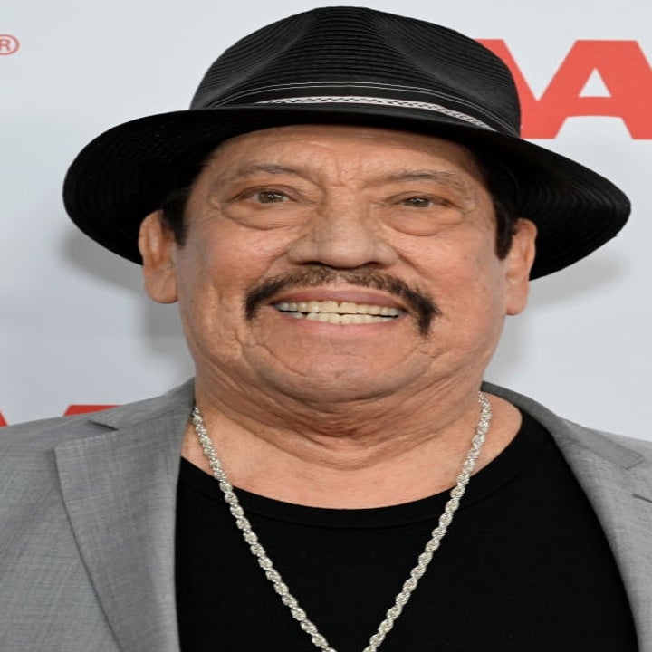 Man in a gray suit, black hat, and chain necklace smiling on an AARP event red carpet