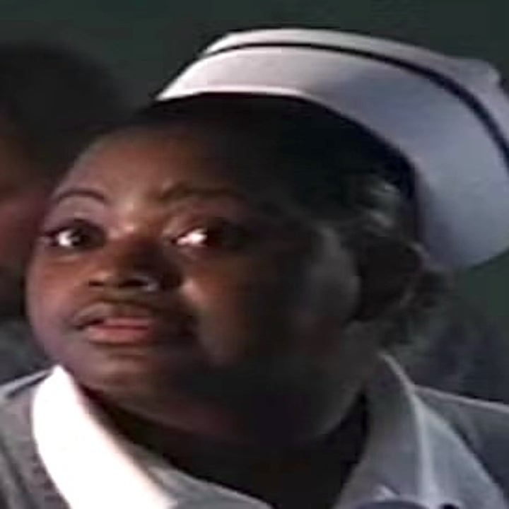 Person wearing a nurse's uniform, looking to the side. Background is blurred, another person is visible