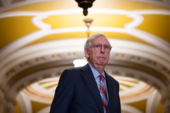 An older man in a suit and striped tie stands in a grand, ornate hallway under an intricate ceiling