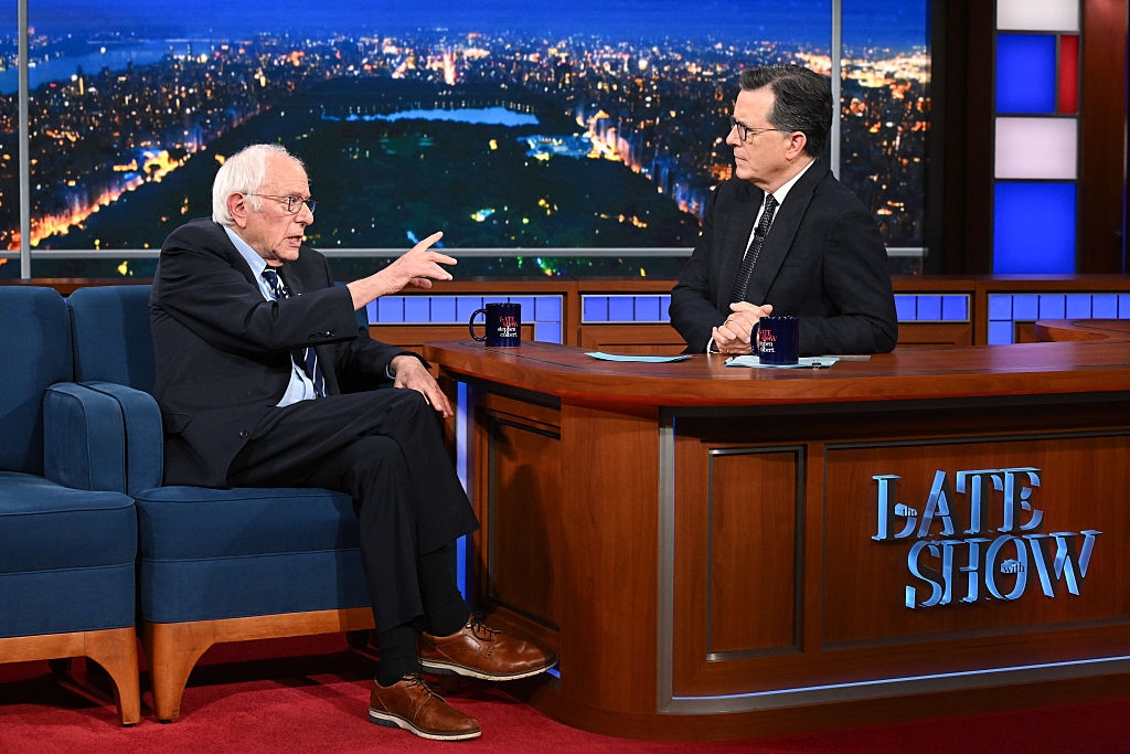 A guest in a suit gestures while speaking to the host seated at a desk on a late-night talk show set with a cityscape backdrop